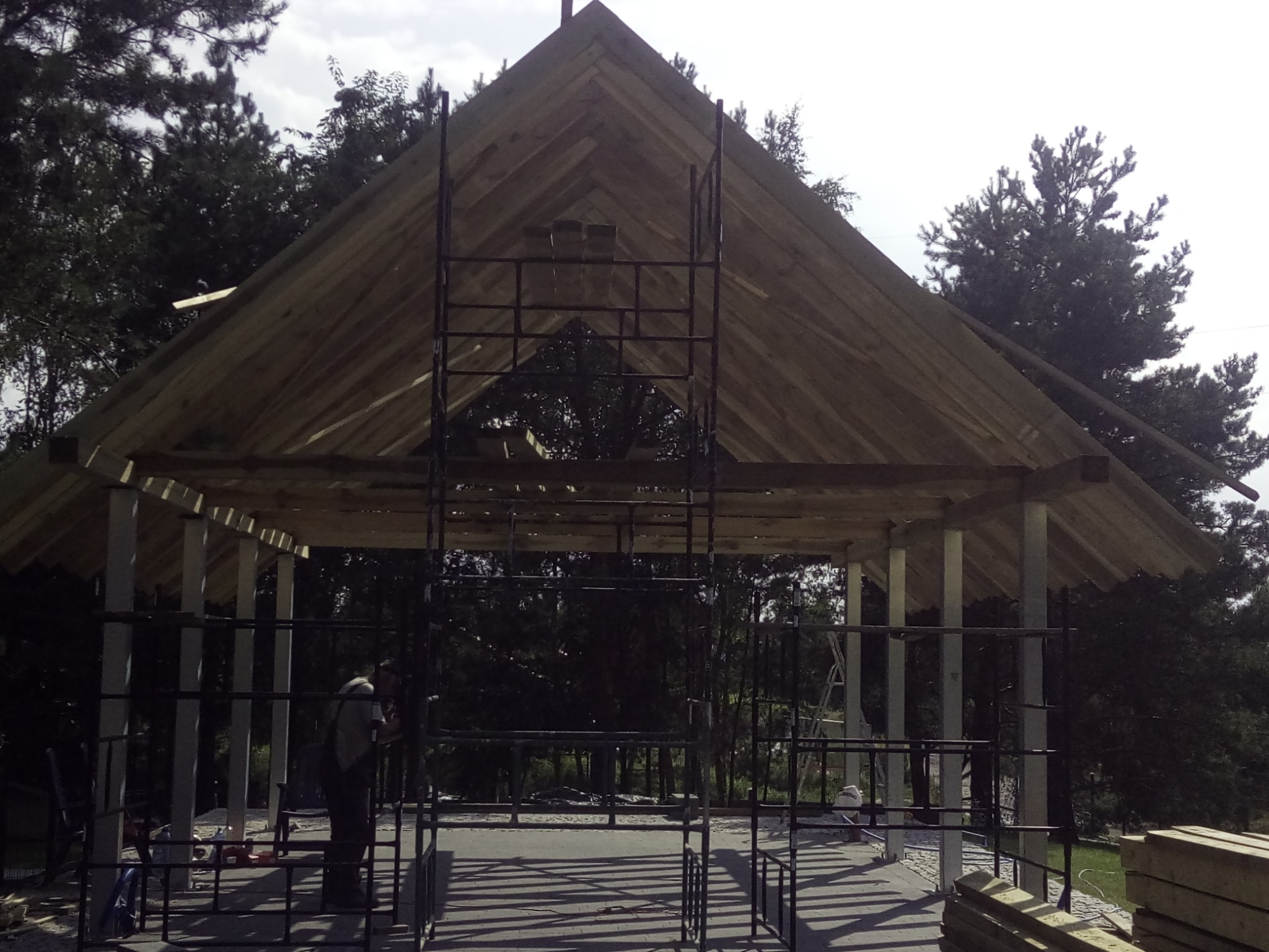 Wznoszenie drewnianej konstrukcji dachu altany z widoczną więźbą, rusztowaniami i częściowo ułożonym pokryciem. W tle zieleń drzew.