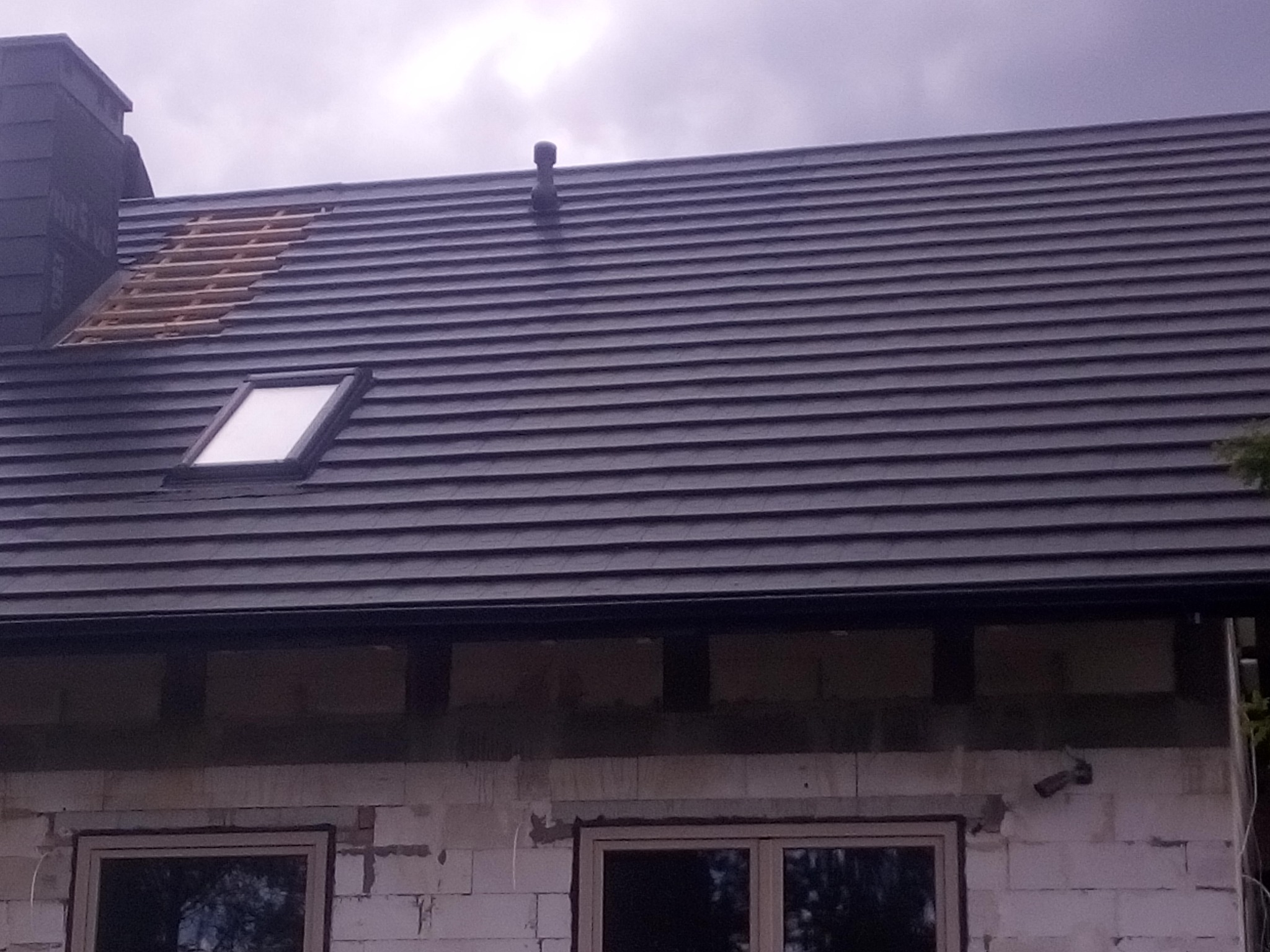 Częściowo pokryty dach z oknem dachowym i fragmentem odsłoniętej więźby, widoczny komin i wentylacja. Budynek w trakcie budowy, ściany z białego pustaka.