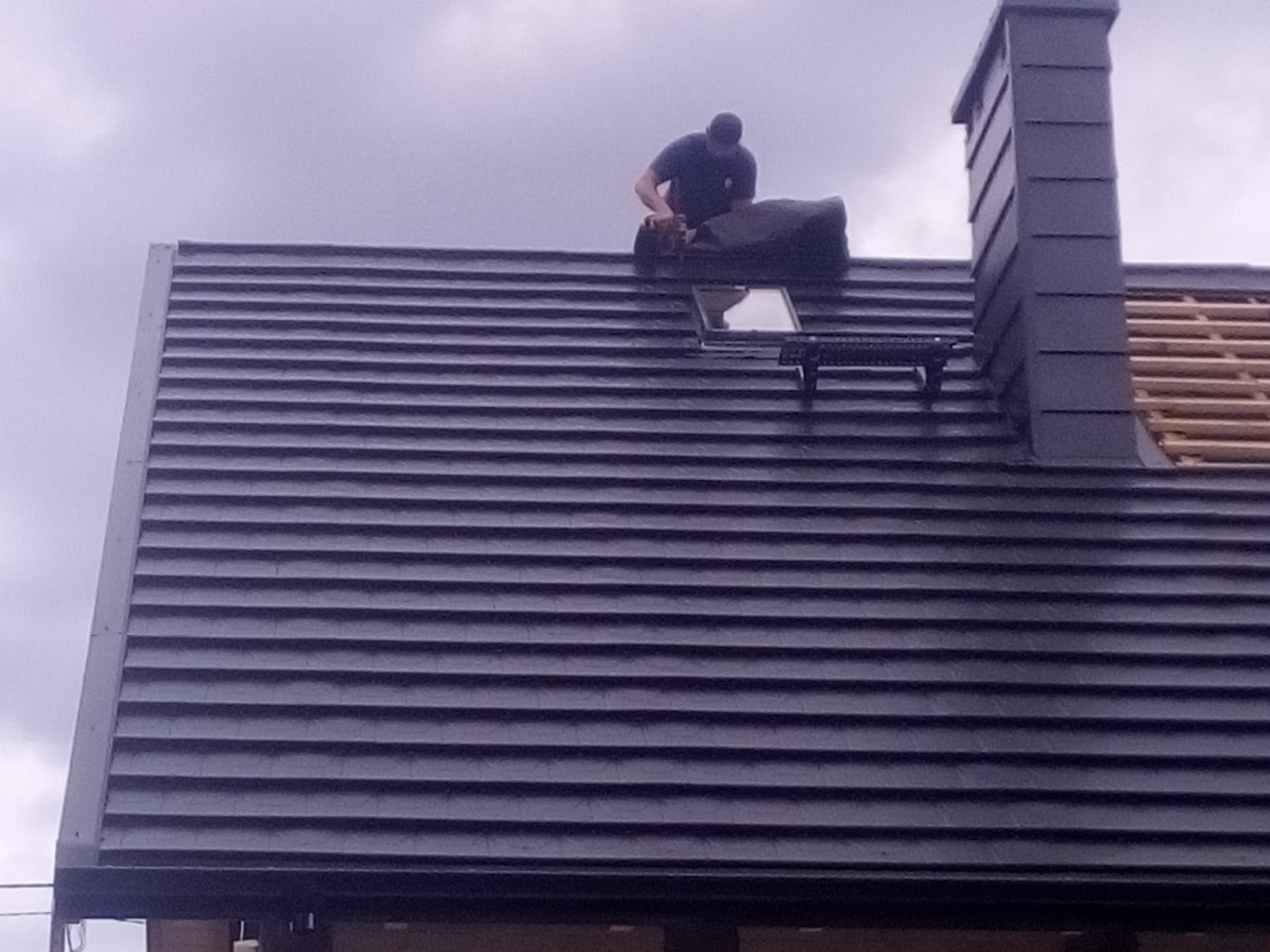 Praca na dachu z ciemnej dachówki, widoczny komin i okno dachowe. Dekarz w trakcie montażu, pochmurne niebo w tle. Częściowo widoczna więźba dachowa.