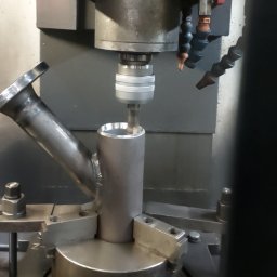 Heisen CNC - Frezarka CNC w trakcie obróbki metalowego elementu z widocznym chłodziwem, zamocowanego w uchwycie. Detal z widocznym spawem i kołnierzem.