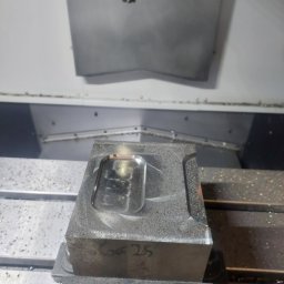 Heisen CNC - Metalowy blok z wyfrezowanym wgłębieniem, zamocowany w uchwycie maszyny CNC. Widoczne wióry i narzędzie skrawające. Napis 'GG 25' na boku bloku.