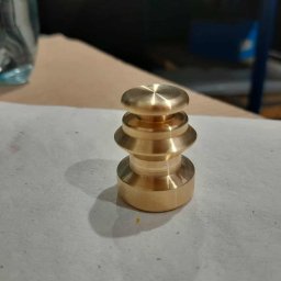 Heisen CNC - Mosiężny element tokarski z trzema pierścieniami, leżący na szarej powierzchni. Widoczne ślady obróbki i odbicia światła. W tle butelka wody i fragment warsztatu.