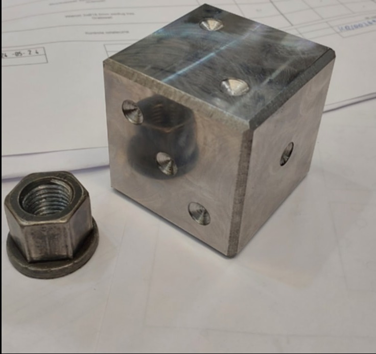 Metalowy sześcian z otworami i nakrętka na tle dokumentów. Widoczne ślady obróbki skrawaniem, ostre krawędzie. Precyzyjne wykonanie detali.