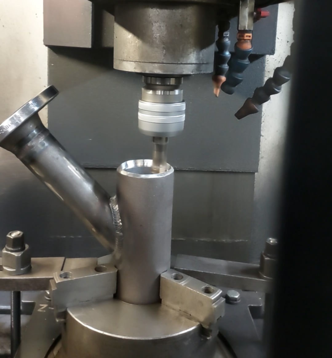 Frezarka CNC w trakcie obróbki metalowego elementu z widocznym chłodziwem, zamocowanego w uchwycie. Detal z widocznym spawem i kołnierzem.