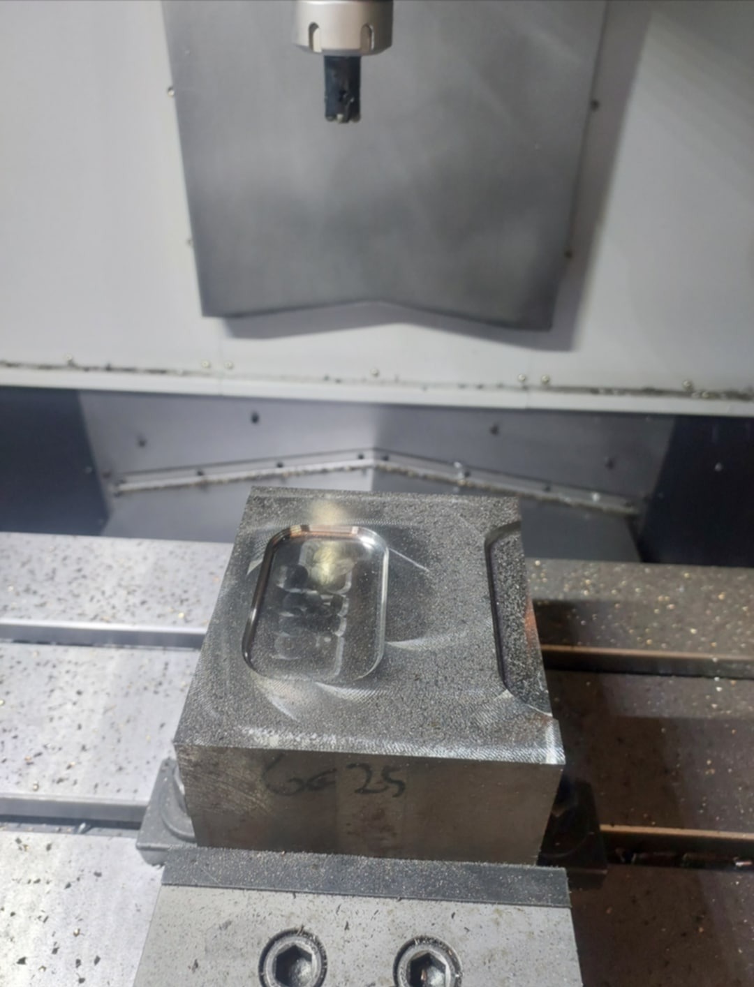 Metalowy blok z wyfrezowanym wgłębieniem, zamocowany w uchwycie maszyny CNC. Widoczne wióry i narzędzie skrawające. Napis 'GG 25' na boku bloku.