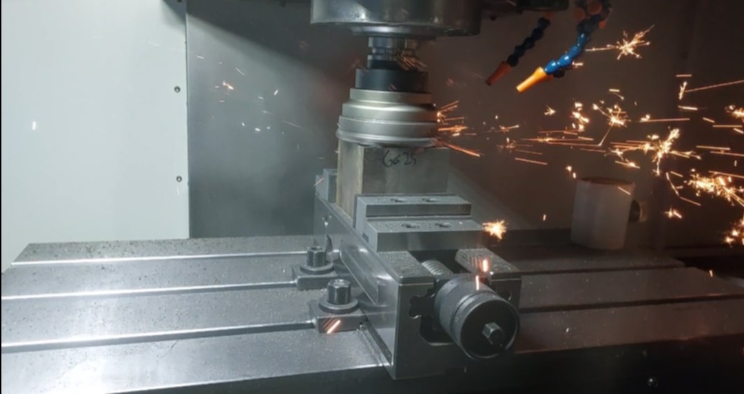 Metalowy blok obrabiany na frezarce CNC, iskry w powietrzu, widoczne dysze chłodzące i uchwyt mocujący. Obróbka skrawaniem w toku.