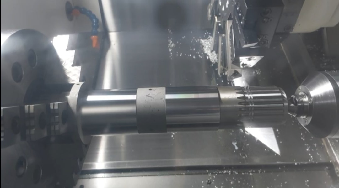 Metalowy wał obrabiany na tokarce CNC, wióry i chłodziwo w trakcie pracy. Widoczne uchwyty tokarki i narzędzie skrawające.