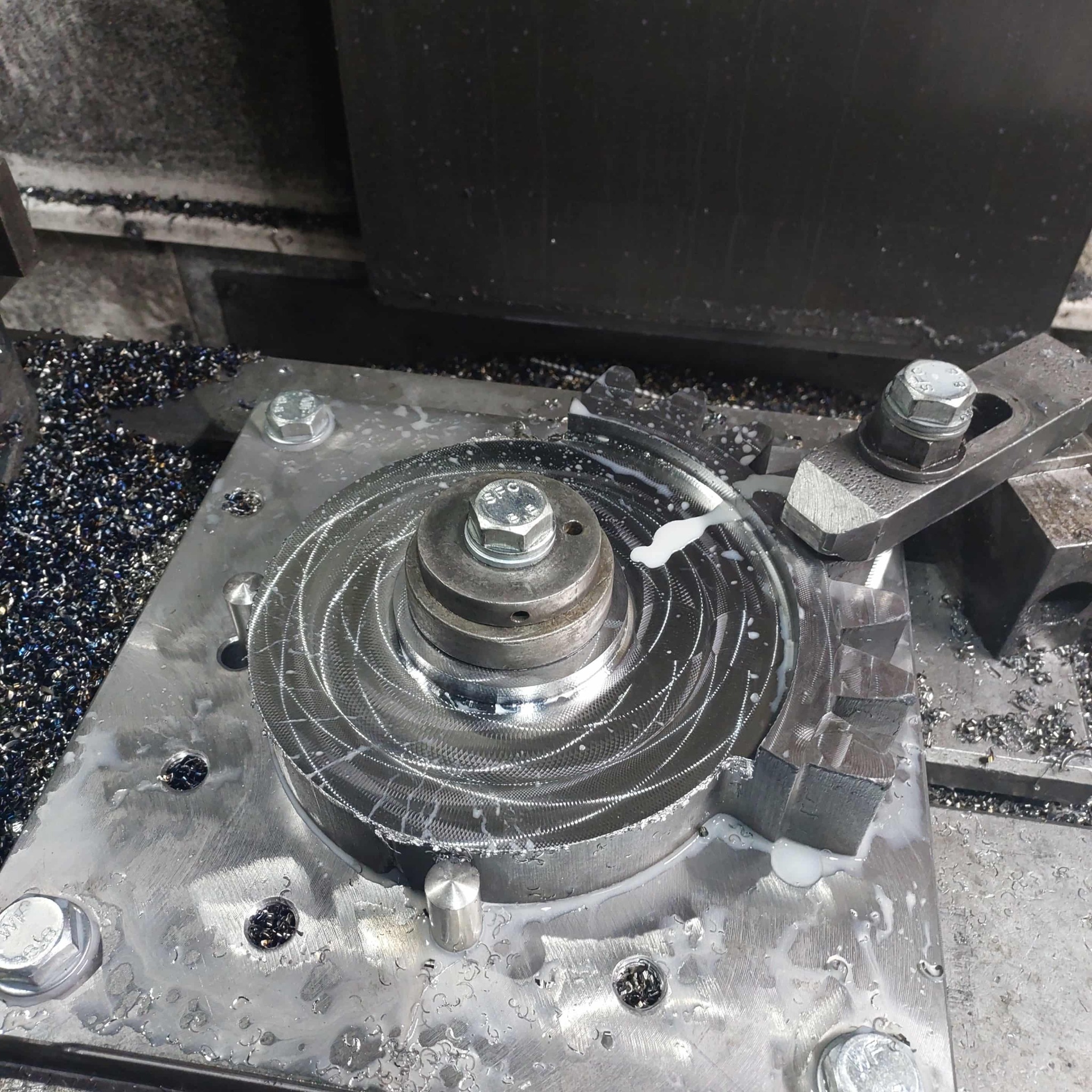 Obrabiany element z metalu na stole frezarki CNC, widoczne wióry i chłodziwo. Detal z wyfrezowanymi rowkami, zamocowany śrubą. Precyzyjna obróbka skrawaniem.