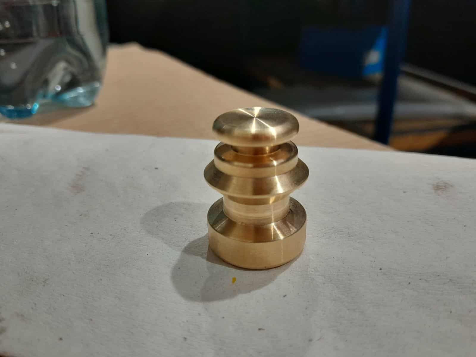 Mosiężny element tokarski z trzema pierścieniami, leżący na szarej powierzchni. Widoczne ślady obróbki i odbicia światła. W tle butelka wody i fragment warsztatu.