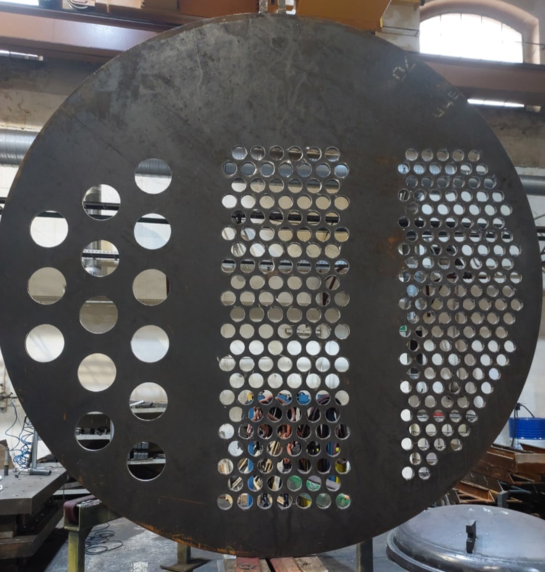 Okrągły, metalowy element z precyzyjnie wyciętymi otworami o różnych średnicach, leżący na warsztatowym stole. Widoczne ślady obróbki i surowy charakter metalu.