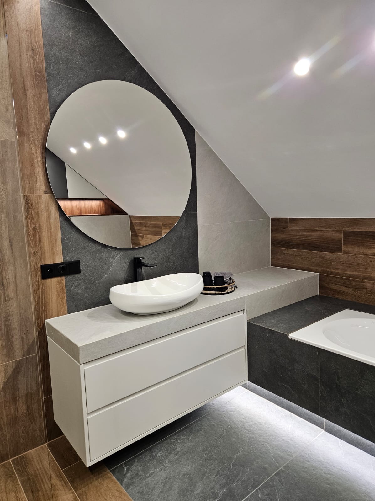Nowoczesna łazienka na poddaszu z owalną umywalką, okrągłym lustrem i podświetleniem LED pod szafką. Ciemne płytki kontrastują z drewnopodobnymi panelami.