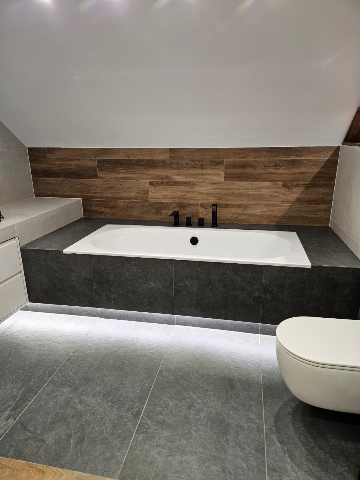 Nowoczesna łazienka na poddaszu z wanną obudowaną ciemnymi płytkami, podświetleniem LED i drewnopodobną okładziną ścienną. Minimalistyczna armatura.