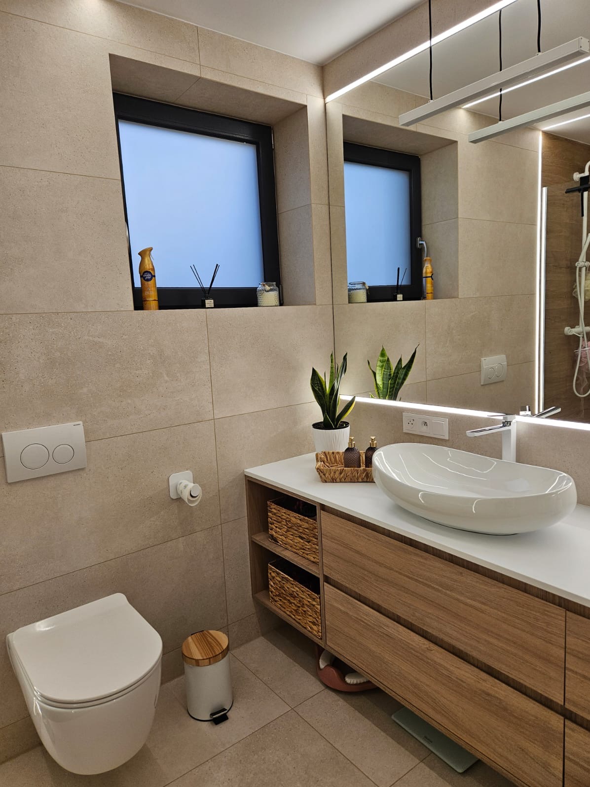Nowoczesna łazienka z beżowymi płytkami, podwieszaną toaletą i umywalką nablatową. Dwa okna z czarnymi ramami i dekoracjami. Oświetlenie LED podkreśla lustro i sufit.