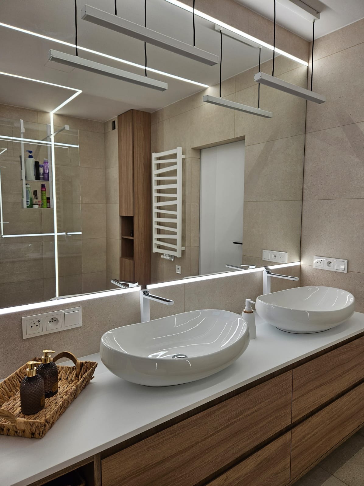 Nowoczesna łazienka z dwoma owalnymi umywalkami na drewnianej szafce, podświetlane lustro z szafką i grzejnikiem w tle. Beżowe płytki na ścianach.