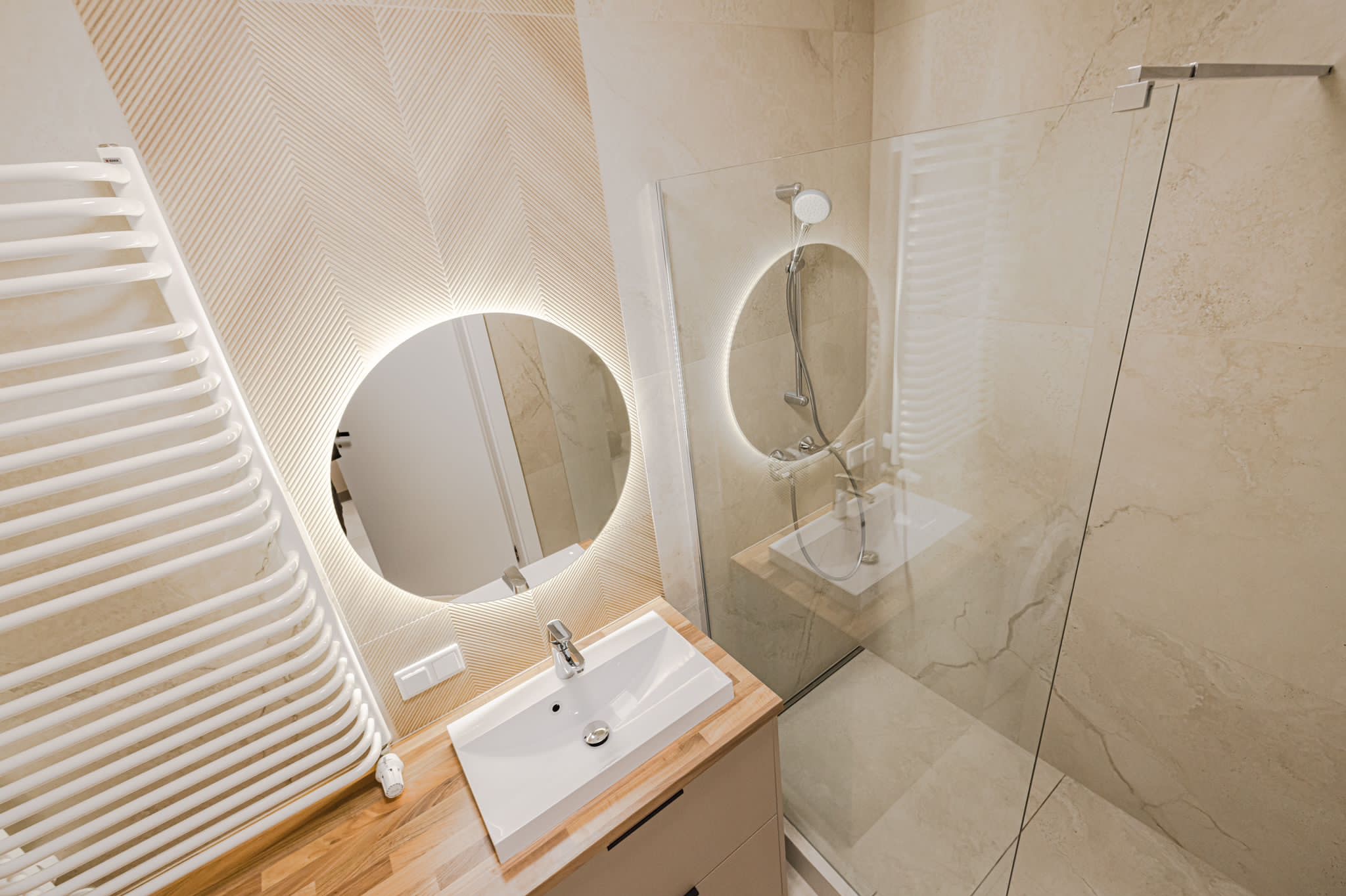 Jasna łazienka z okrągłym lustrem LED, kabiną prysznicową ze szkła i grzejnikiem drabinkowym. Umywalka nablatowa na drewnianym blacie. Ściany wyłożone płytkami.
