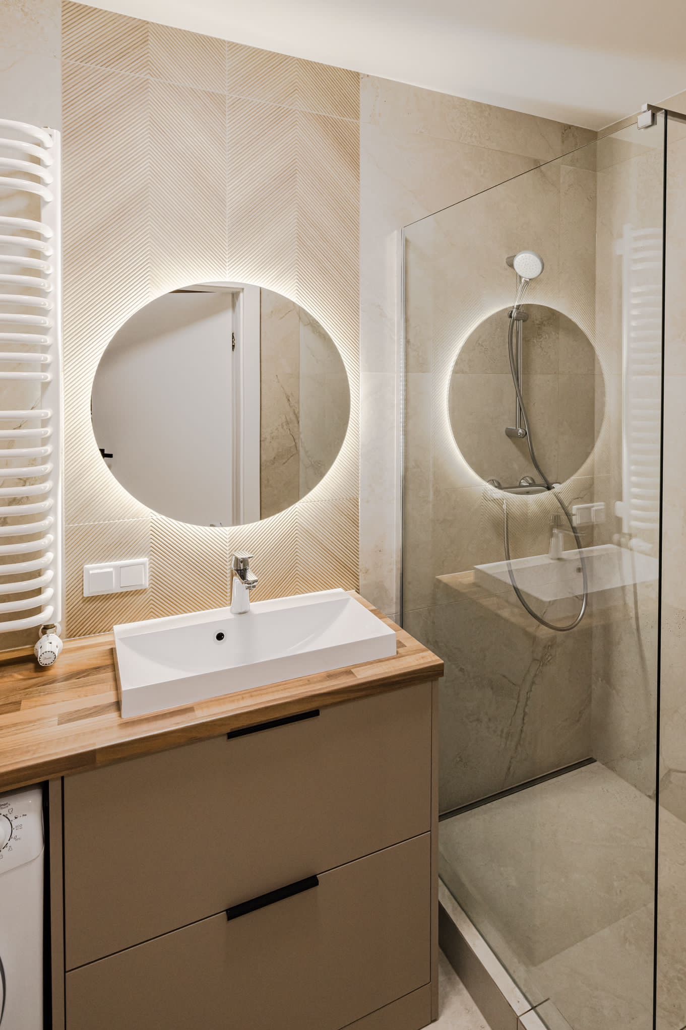Nowoczesna łazienka z okrągłym lustrem z podświetleniem LED, kabiną prysznicową ze szkła i umywalką na szafce z drewnianym blatem. Minimalistyczny design.