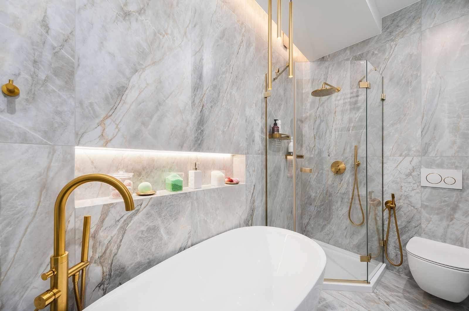 Elegancka łazienka w Trzemeśni z marmurowymi płytkami, złotą armaturą i kabiną prysznicową. Nowoczesny design z podświetlaną wnęką i wolnostojącą wanną.