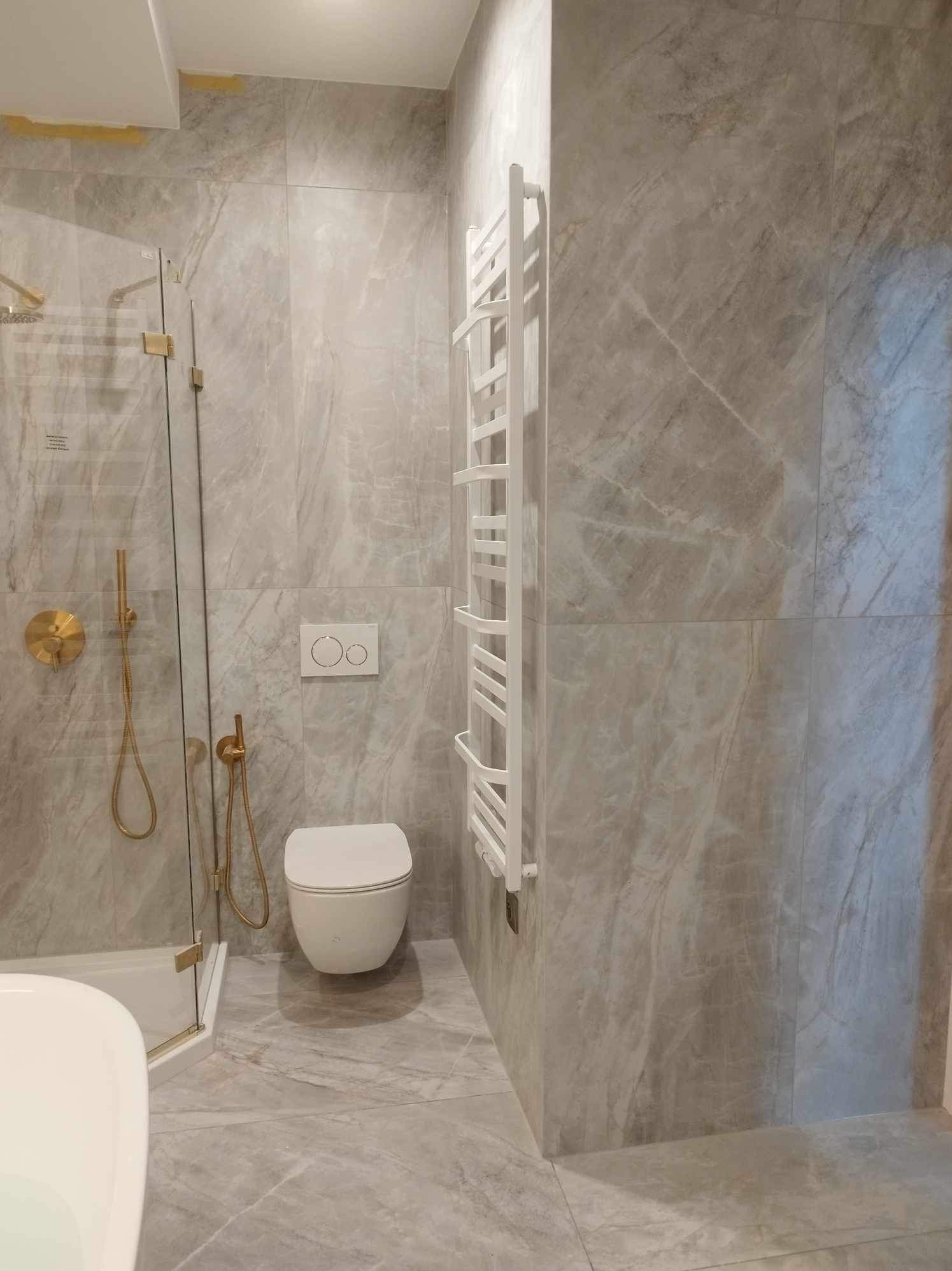 Wykończona łazienka z marmurowymi płytkami, złotą armaturą prysznicową i białym grzejnikiem drabinkowym. Minimalistyczny design, widok na kabinę i toaletę.