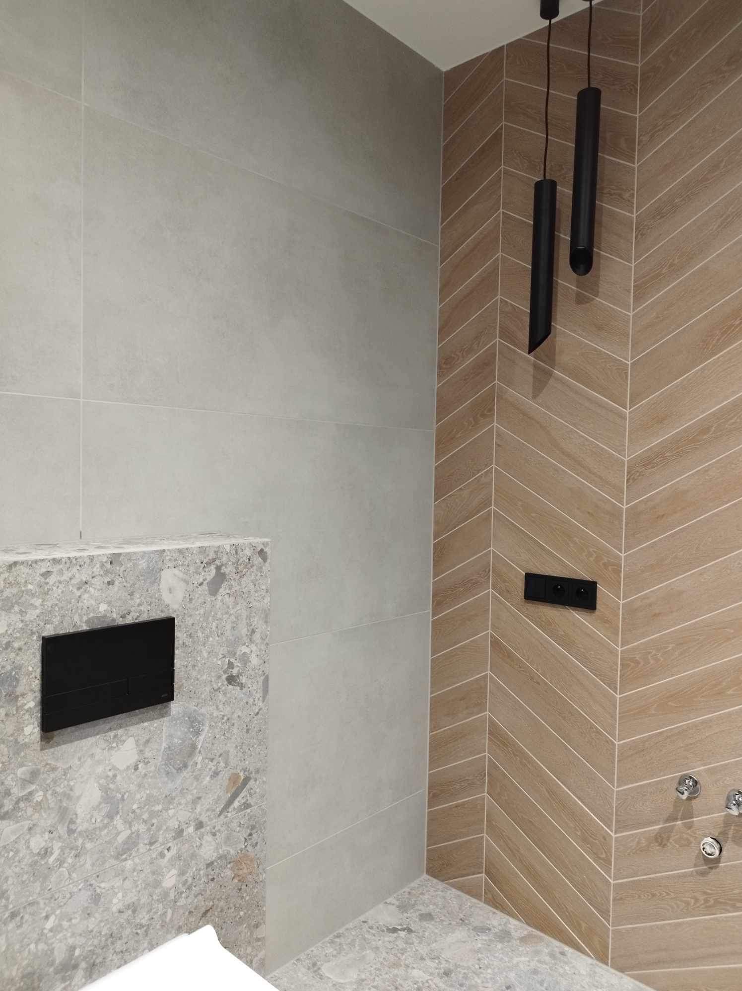 Nowoczesna łazienka z płytkami imitującymi drewno ułożonymi w jodełkę, szare płytki i marmurowa zabudowa WC z czarnym przyciskiem. Minimalistyczne lampy.