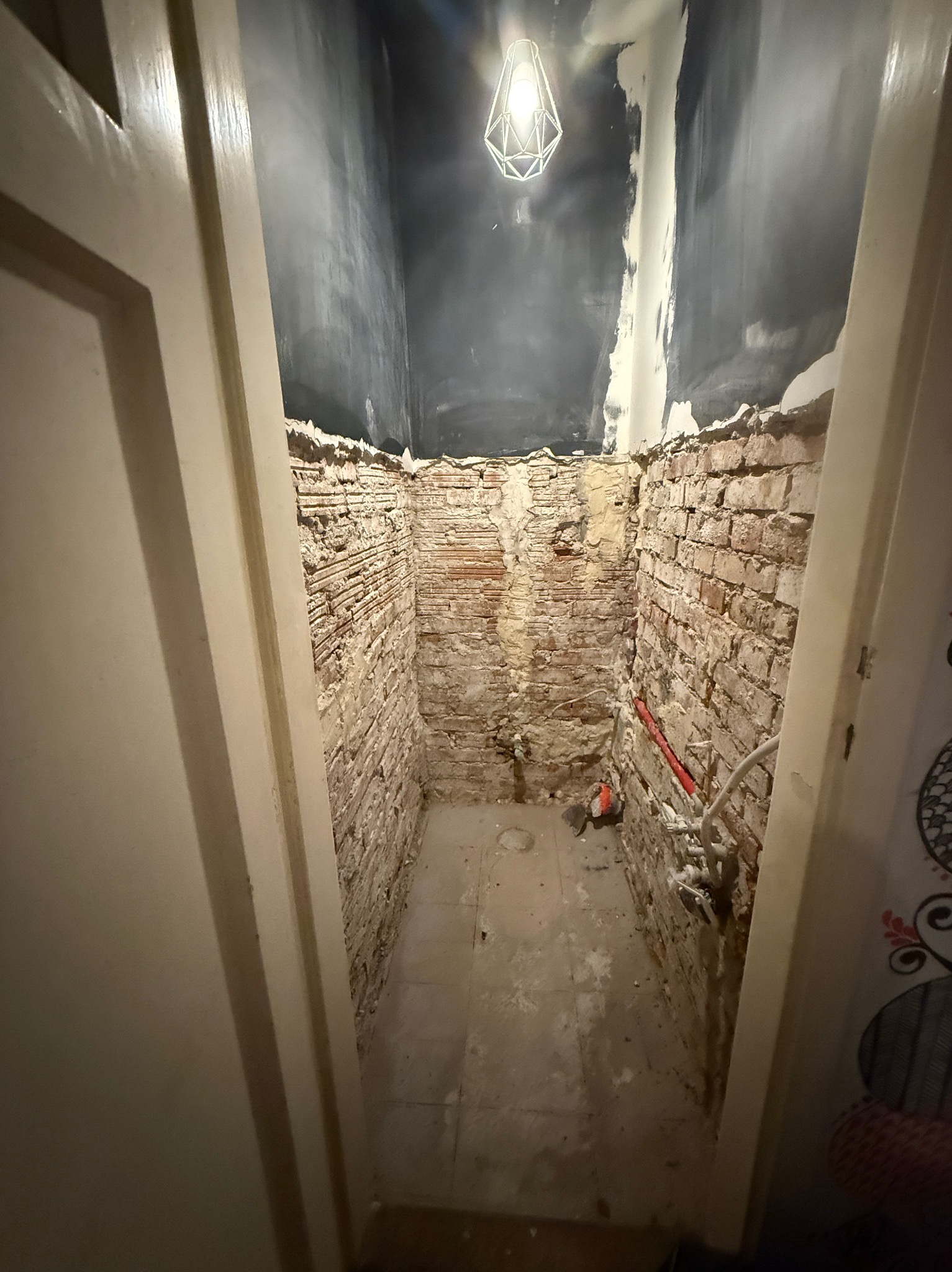 Wnętrze małej łazienki w trakcie remontu. Odsłonięte cegły na ścianach, widoczne instalacje, surowy stan przed wykończeniem. Wisząca lampa oświetla przestrzeń.