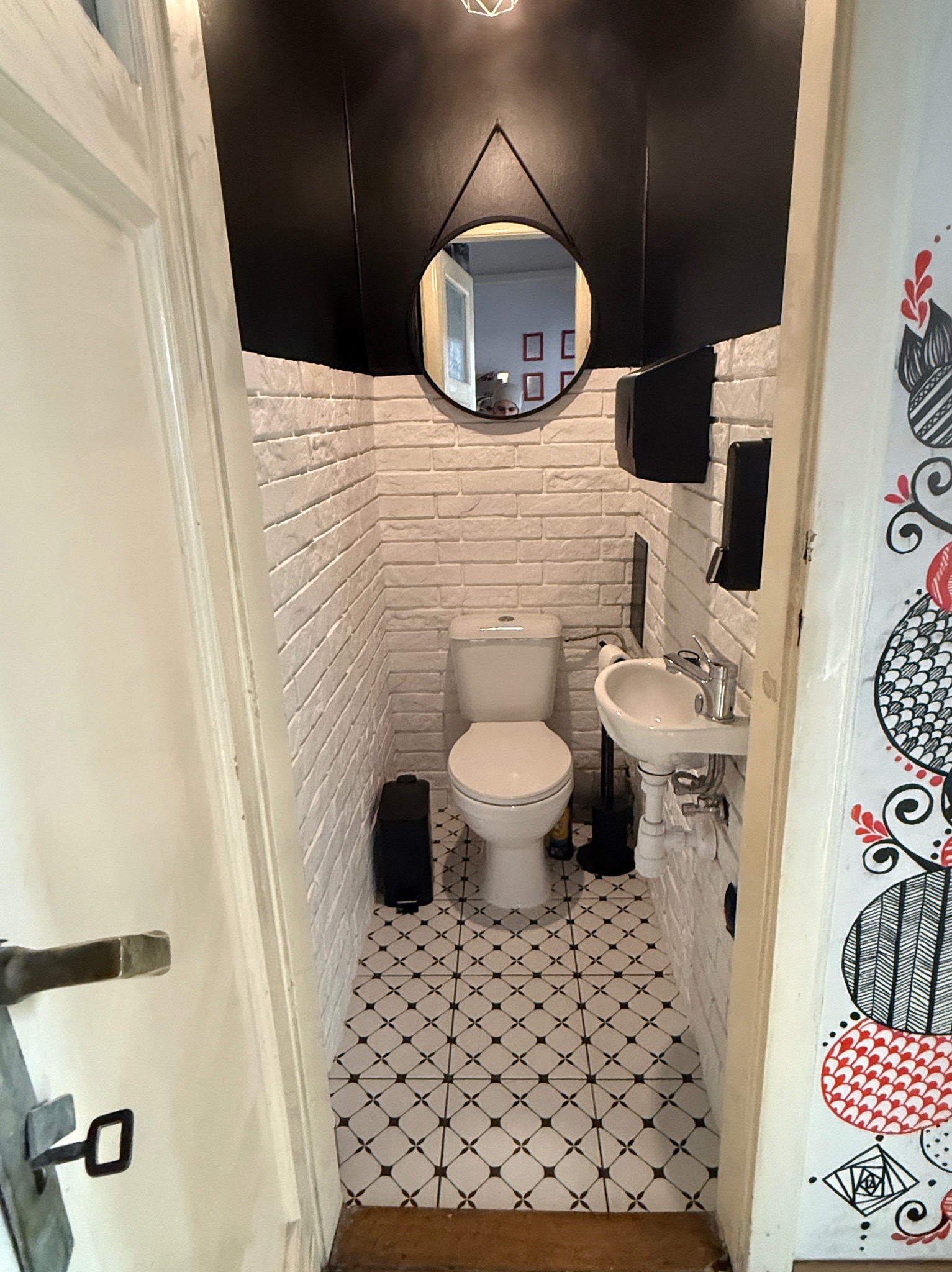 Wąska łazienka z białą cegłą, czarnym sufitem i wzorzystą podłogą. Okrągłe lustro wisi nad toaletą. Drzwi z dekoracyjnym malunkiem po prawej stronie.