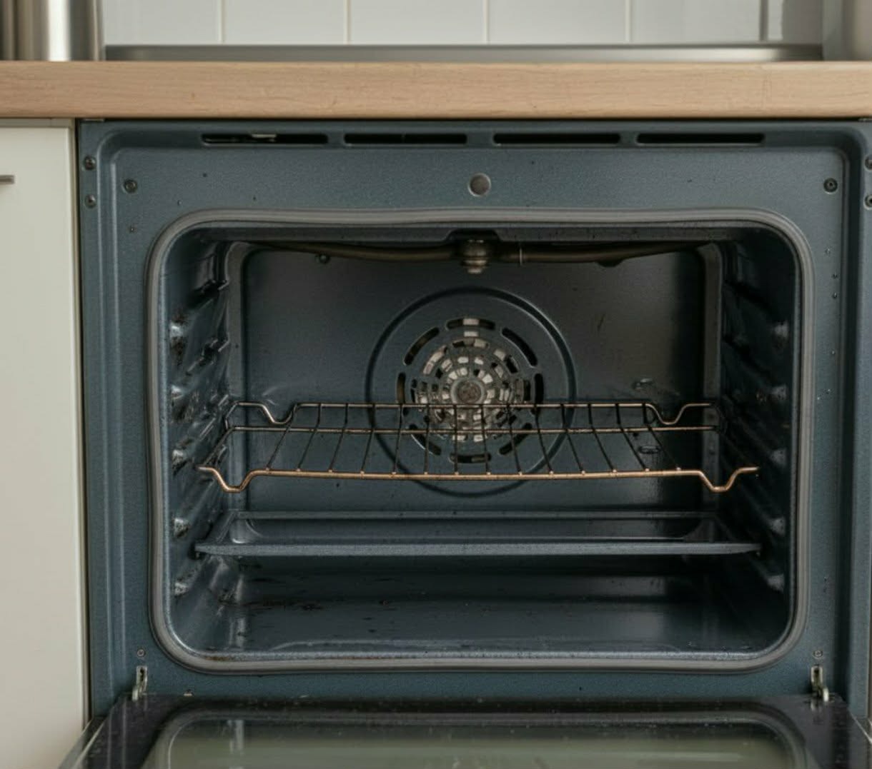 Wnętrze otwartego piekarnika z rusztem i wentylatorem, widoczne ślady użytkowania. Tło stanowią białe płytki i fragment blatu kuchennego.