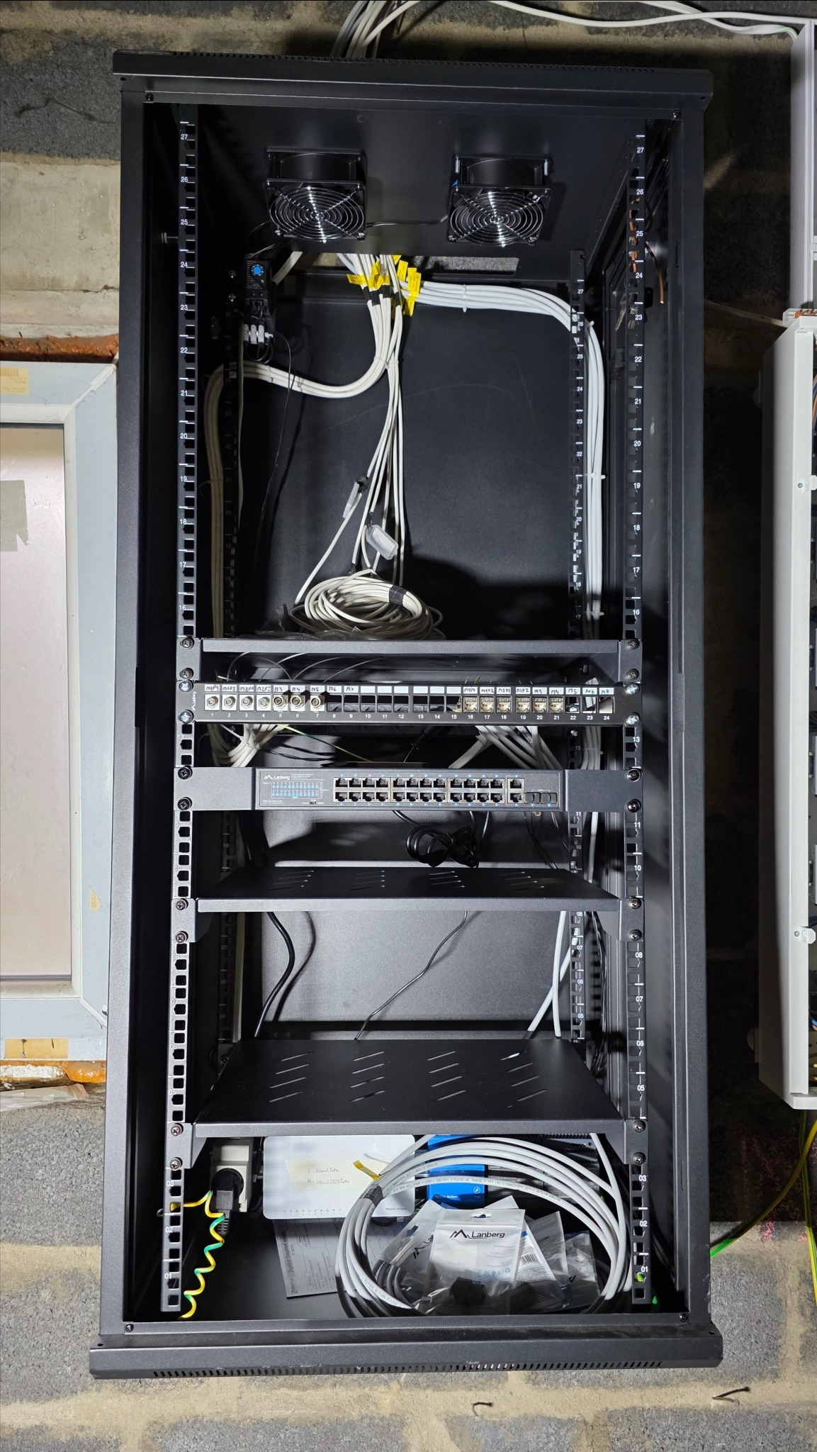 Szafa rack z zamontowanym sprzętem sieciowym: switche, patch panele i okablowanie. Widoczne wentylatory chłodzące i zasilanie. Elementy inteligentnego domu.