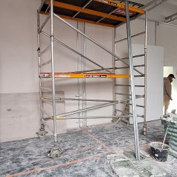 Rem-Bud Waw - Wnętrze w trakcie remontu: metalowe rusztowanie na kółkach, widoczne profile sufitowe, ślady farby na podłodze, osoba w tle. Etap prac wykończeniowych.