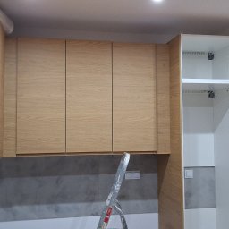 Rem-Bud Waw - Jasne, drewniane szafki kuchenne wiszące nad szarą ścianą, z częściowo zamontowanym białym regałem obok. Widoczna drabina sugeruje trwający montaż.