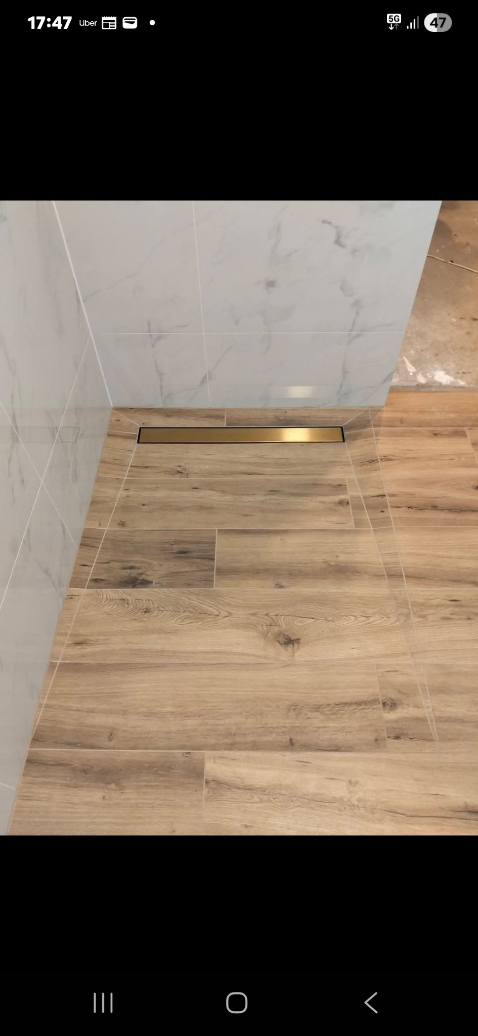 Wykończona łazienka z drewnopodobnymi płytkami podłogowymi i odpływem liniowym w kolorze złotym, połączona z białymi płytkami ściennymi imitującymi marmur.