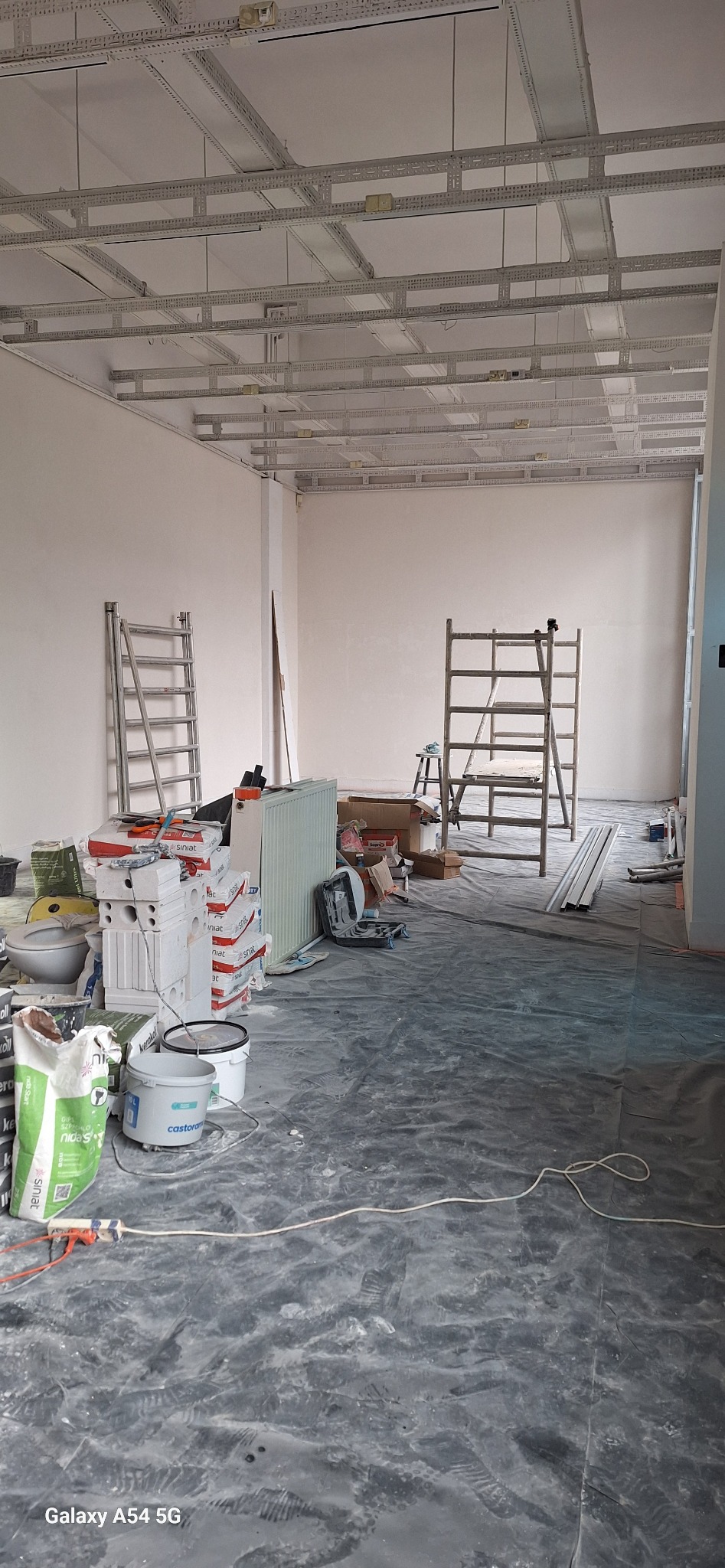Wnętrze w trakcie prac wykończeniowych: widoczne materiały budowlane, drabiny i stelaż sufitu podwieszanego. Podłoga zabezpieczona folią malarską.