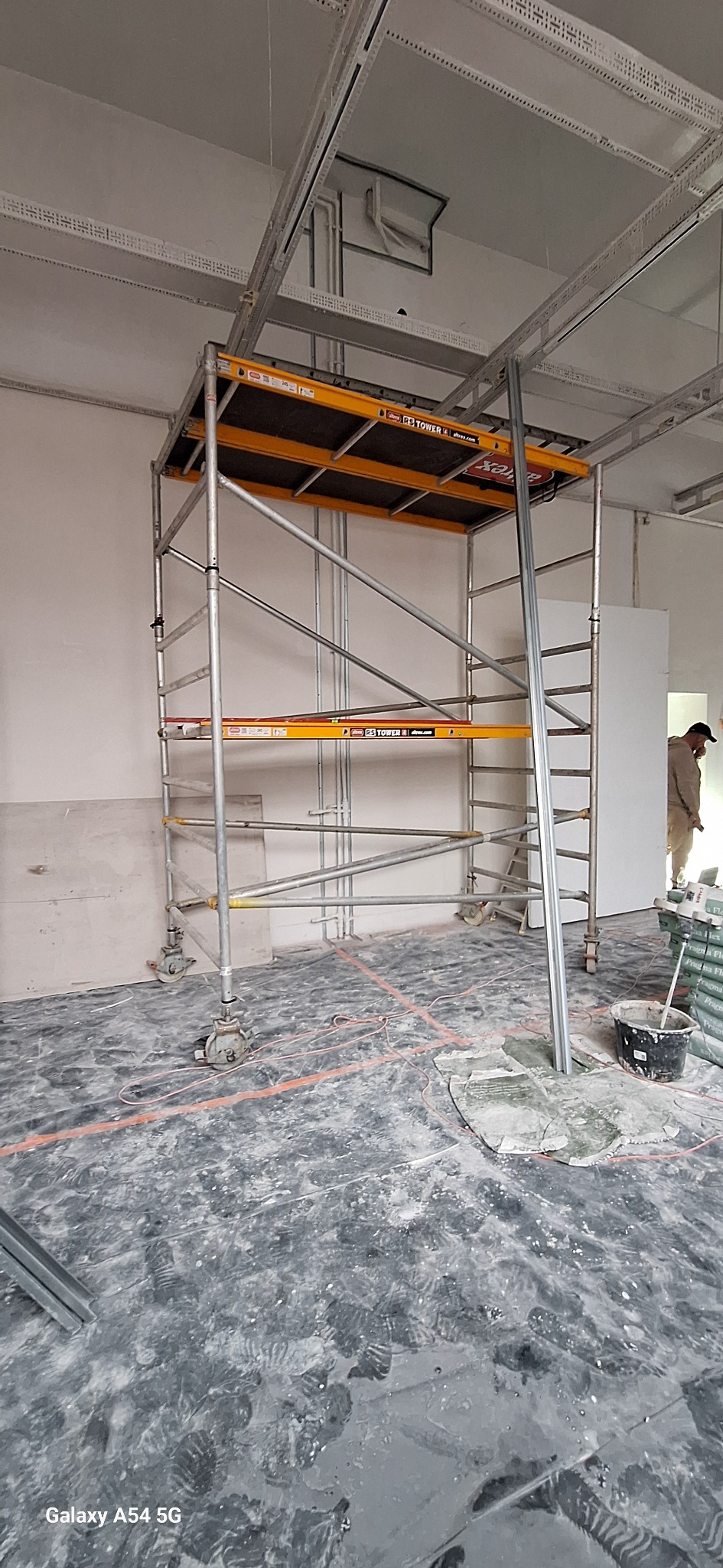 Wnętrze w trakcie remontu: metalowe rusztowanie na kółkach, widoczne profile sufitowe, ślady farby na podłodze, osoba w tle. Etap prac wykończeniowych.