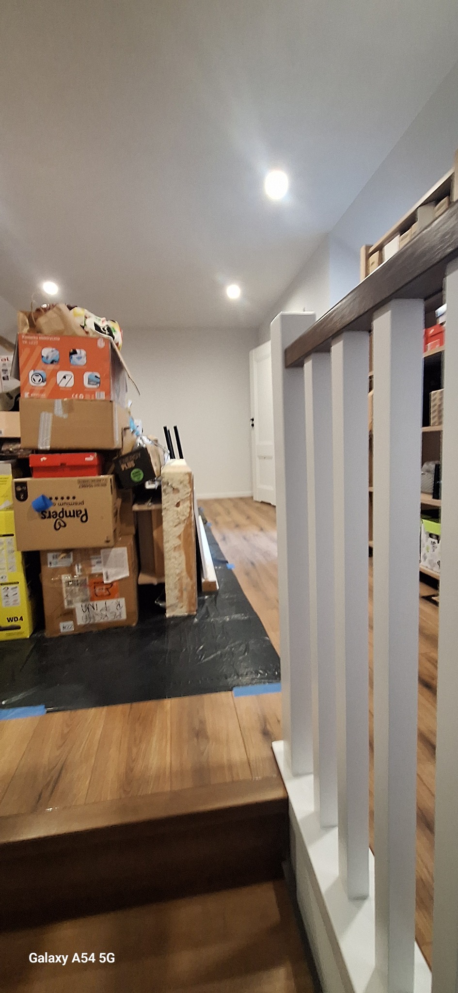 Przygotowania do remontu: podłoga zabezpieczona folią, sterta kartonów i materiałów budowlanych, widoczne elementy balustrady. Wnętrze w trakcie prac.