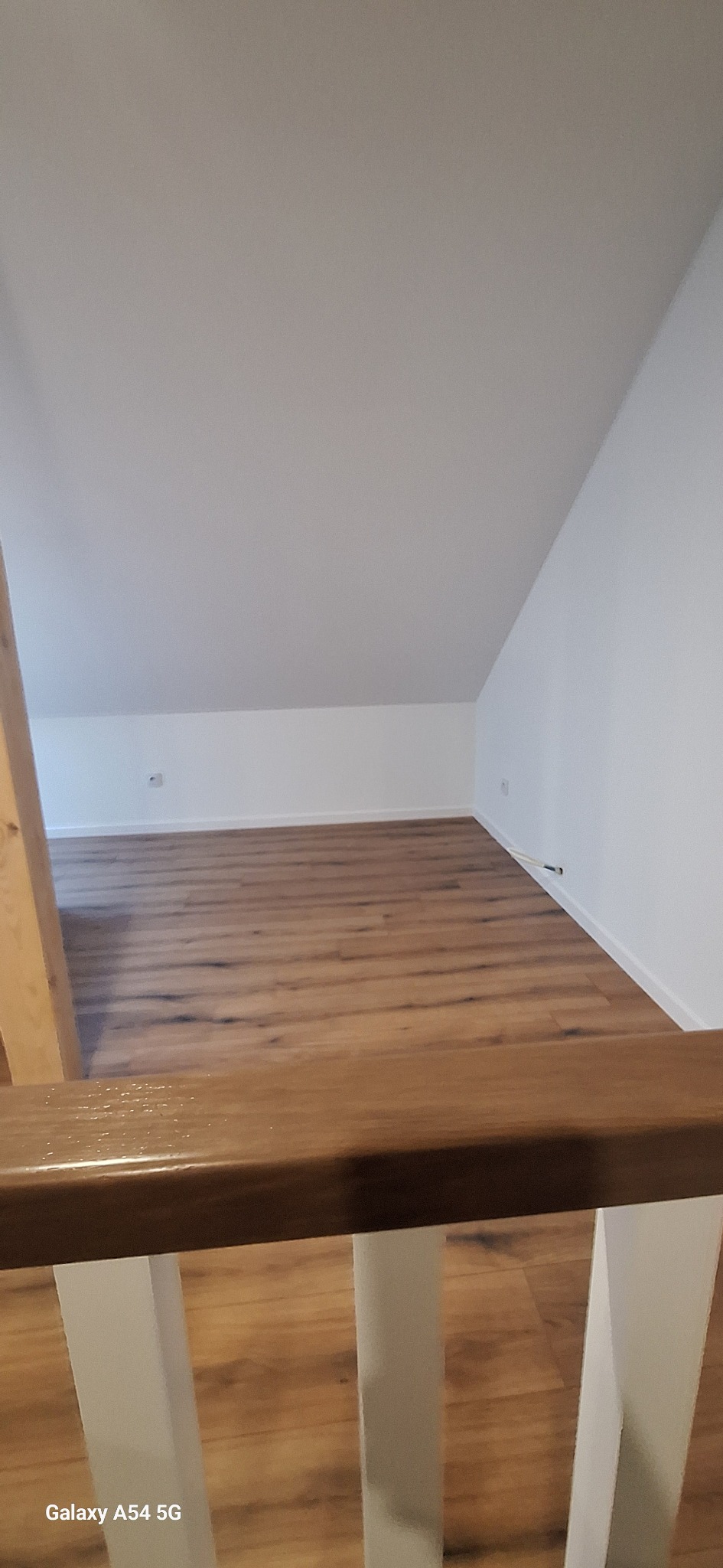 Poddasze z drewnianą podłogą i białymi ścianami, widok z góry przez drewnianą balustradę. Minimalistyczne wnętrze po remoncie, z widocznymi detalami wykończenia.