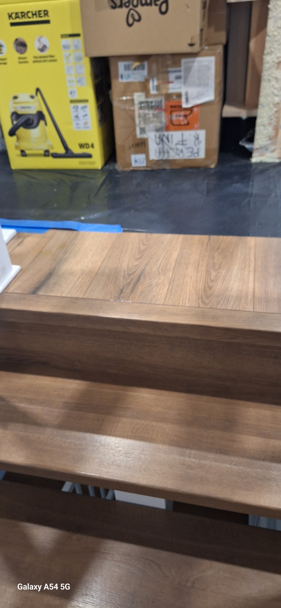 Wykończone drewniane schody z widoczną strukturą drewna, w tle kartony i sprzęt. Ujęcie z dołu, podkreślające detale wykończenia stopni.