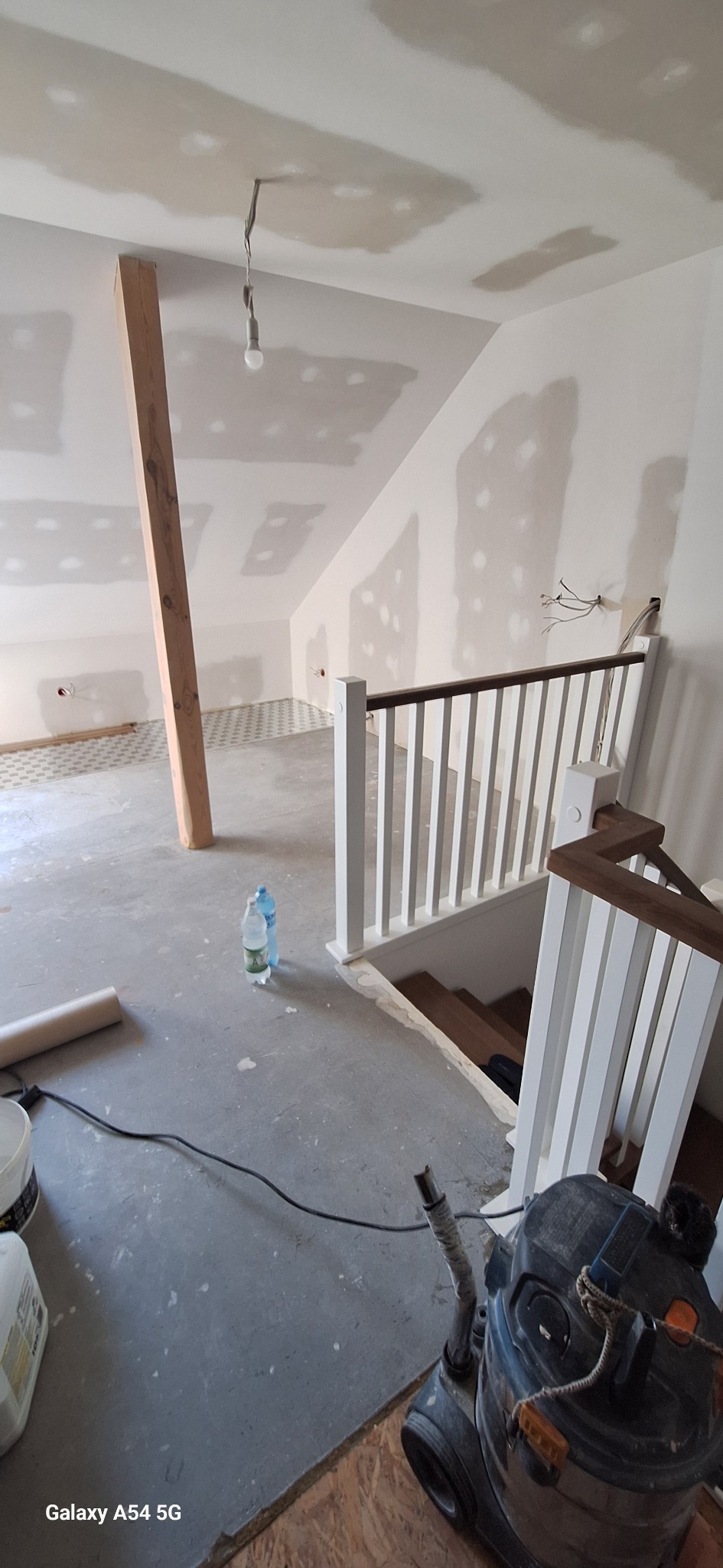 Wnętrze w trakcie wykańczania: surowy beton, częściowo położona podłoga, oświetlenie tymczasowe, schody z balustradą i narzędzia budowlane. Widoczne ślady szpachlowania na ścianach.