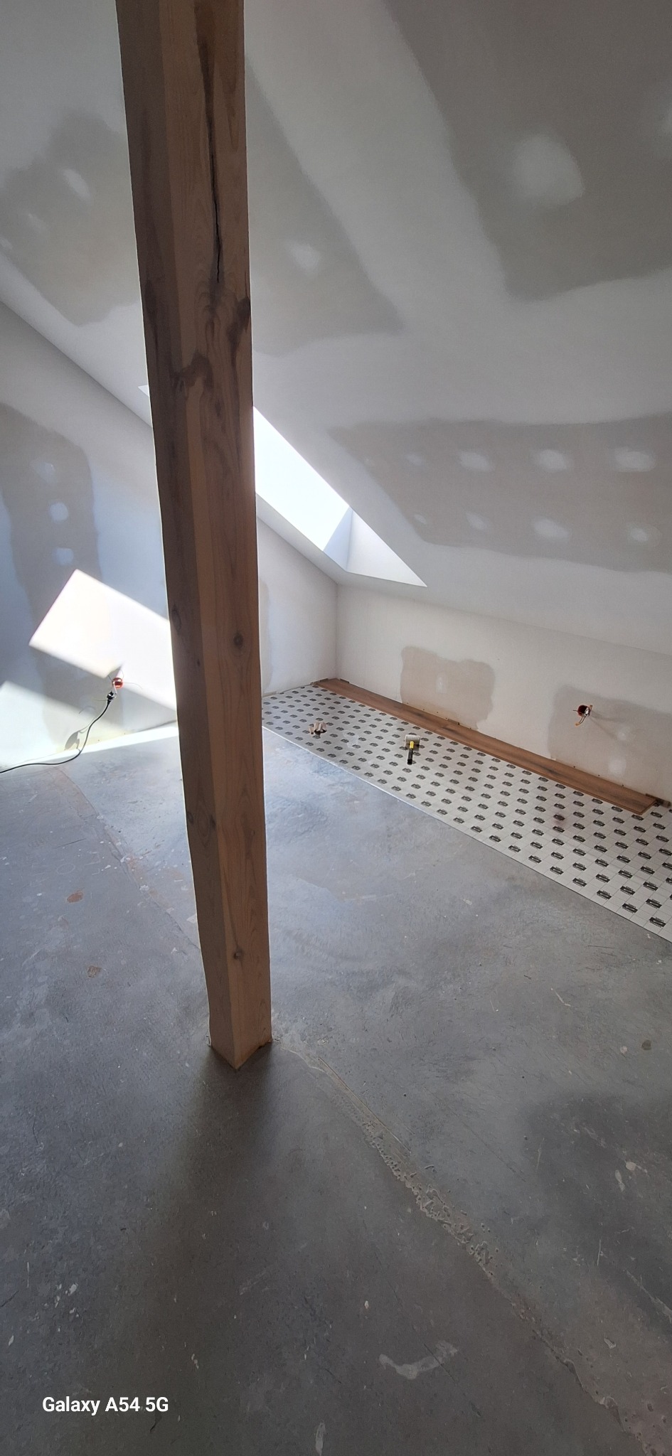 Poddasze w trakcie prac wykończeniowych: drewniany słup, częściowo ułożona podłoga z wzorem, przygotowane ściany do malowania, okno dachowe. Widok na wnętrze w trakcie remontu.