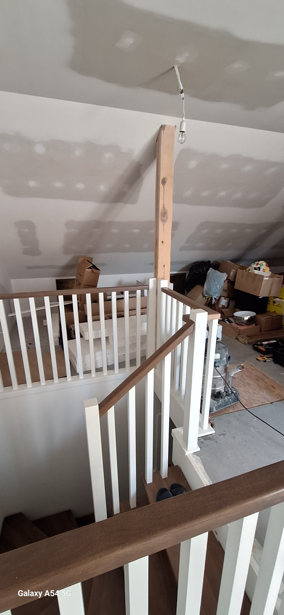 Wnętrze poddasza w trakcie wykańczania: białe balustrady schodów z drewnianymi poręczami, surowy słup podtrzymujący, ściany przygotowane do malowania, kartony w tle.