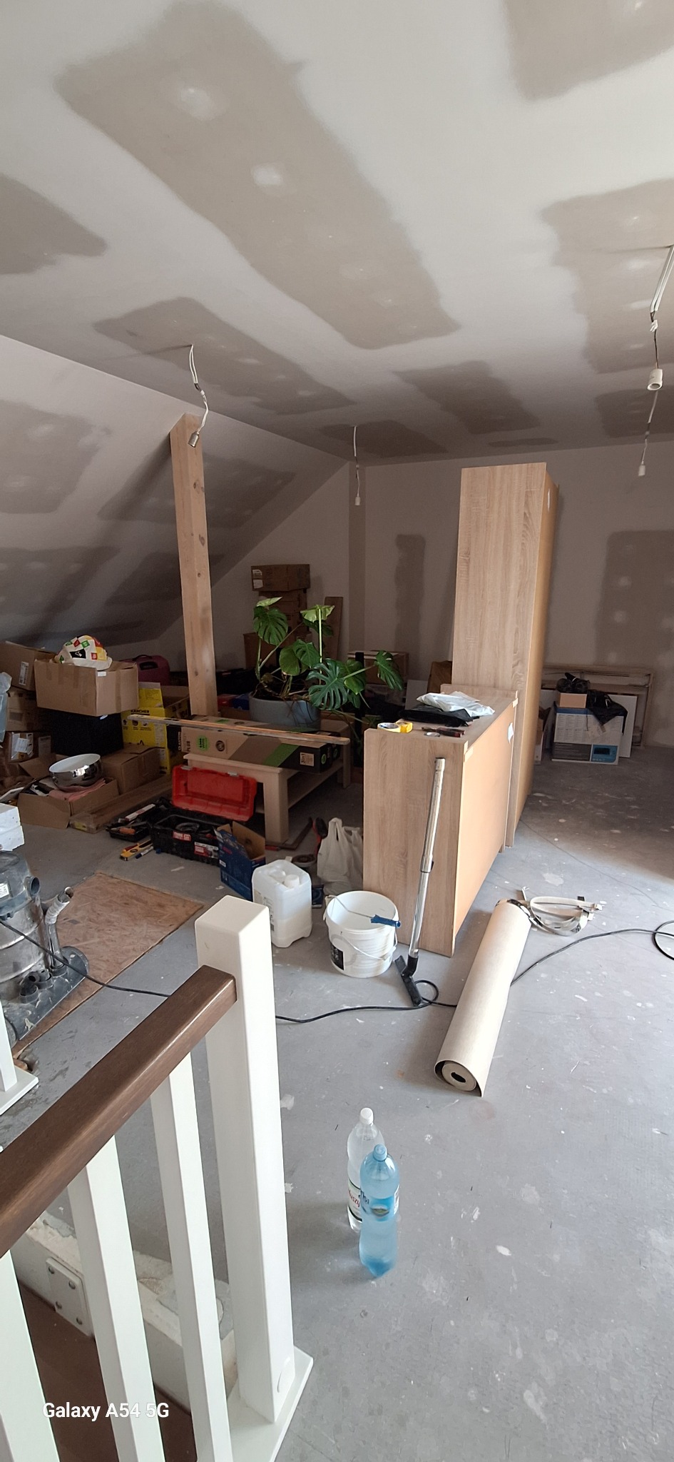 Poddasze w trakcie wykańczania: płyty gipsowe na skosach, widoczne narzędzia, materiały budowlane i elementy wyposażenia wnętrz. Roślina doniczkowa dodaje koloru.