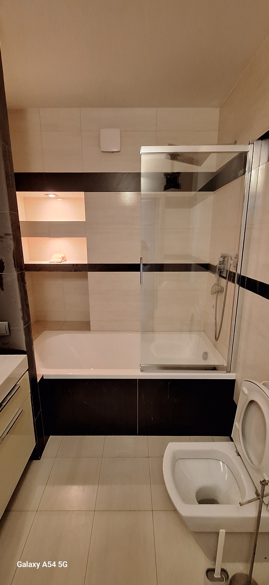 Wykończona łazienka z beżowymi płytkami, czarnymi akcentami i oświetlonymi niszami. Widoczna wanna z parawanem, toaleta i umywalka z szafką. Minimalistyczny design.