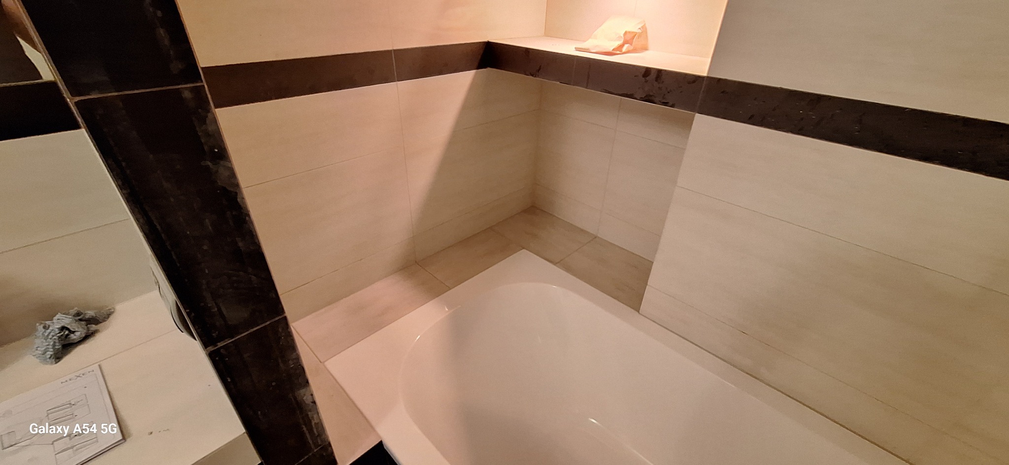 Wykończona łazienka z wanną, jasnymi płytkami imitującymi drewno i ciemnobrązowymi akcentami. Na półce wnęki leży zwinięty ręcznik. Widoczny fragment podłogi i ściany.