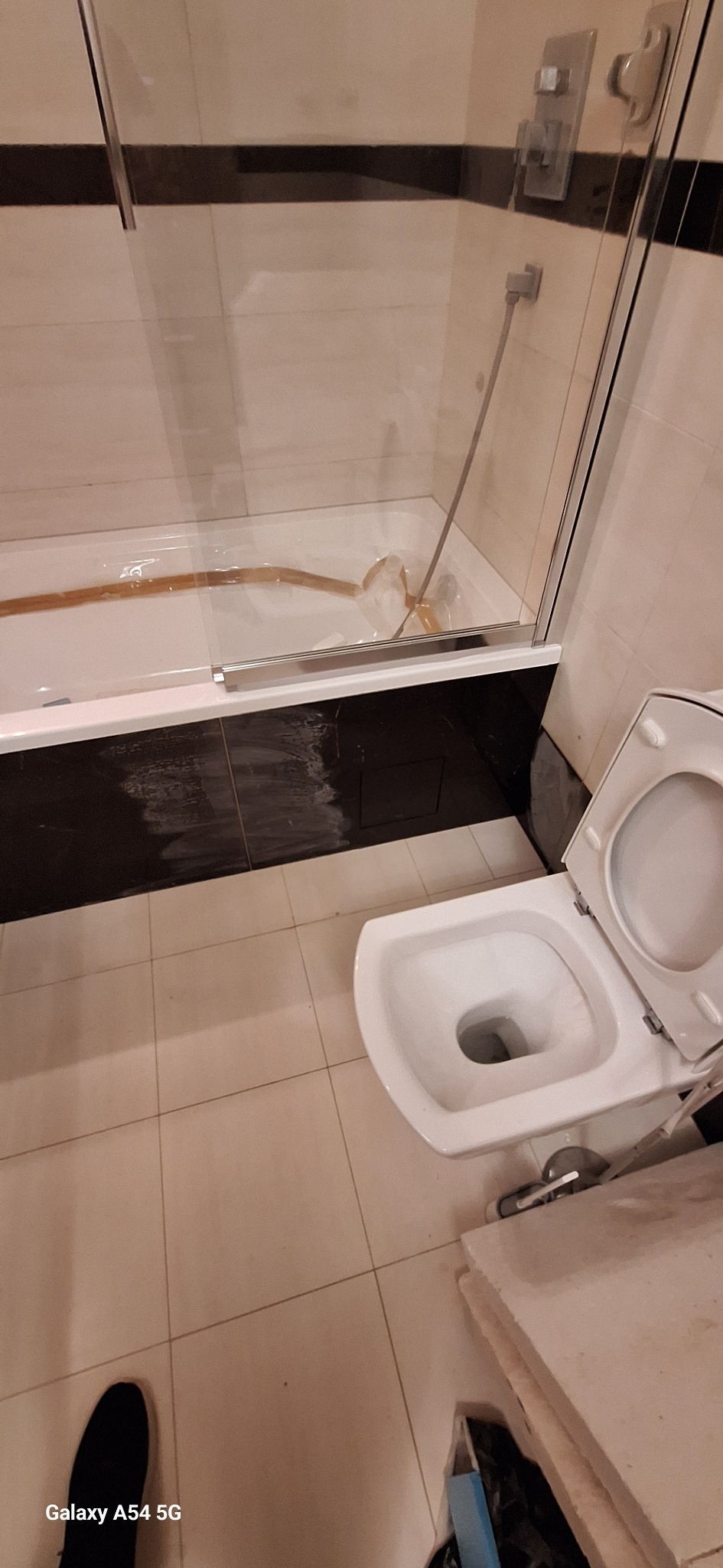 Wykończona łazienka z kabiną prysznicową, wanną i toaletą. Widoczne beżowe płytki podłogowe i czarne płytki na obudowie wanny. Ujęcie z góry, widoczny fragment buta.