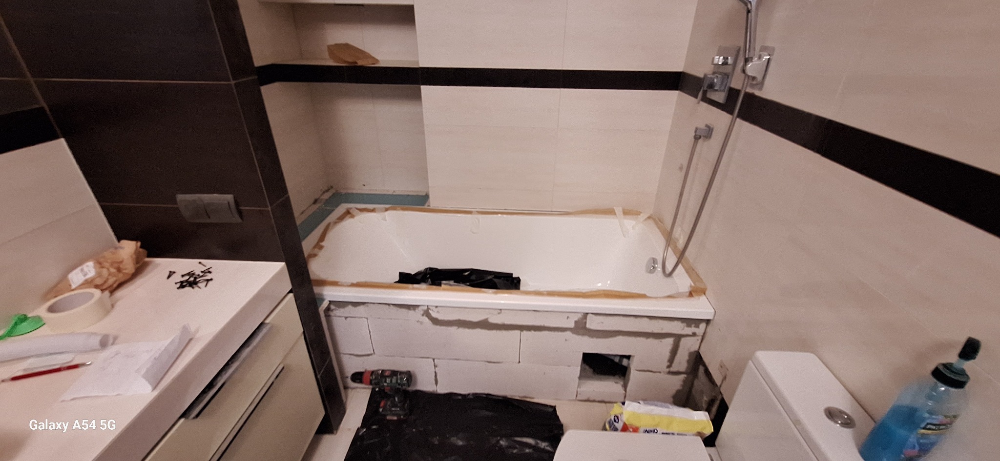 Biała wanna obudowana bloczkami w łazience z czarno-białymi płytkami. Widoczne narzędzia i materiały budowlane. Etap prac wykończeniowych.