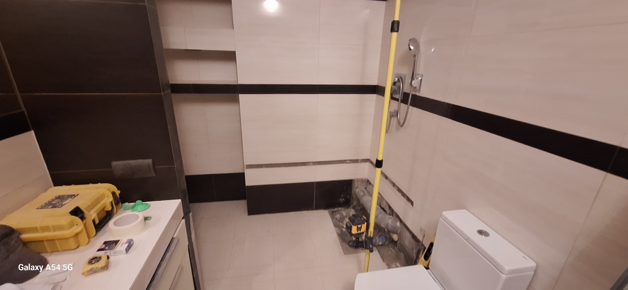 Remont łazienki w trakcie: częściowo wykończone płytki, narzędzia glazurnicze i toaleta. Widoczny laser krzyżowy. Prace wykończeniowe w nowoczesnym stylu.