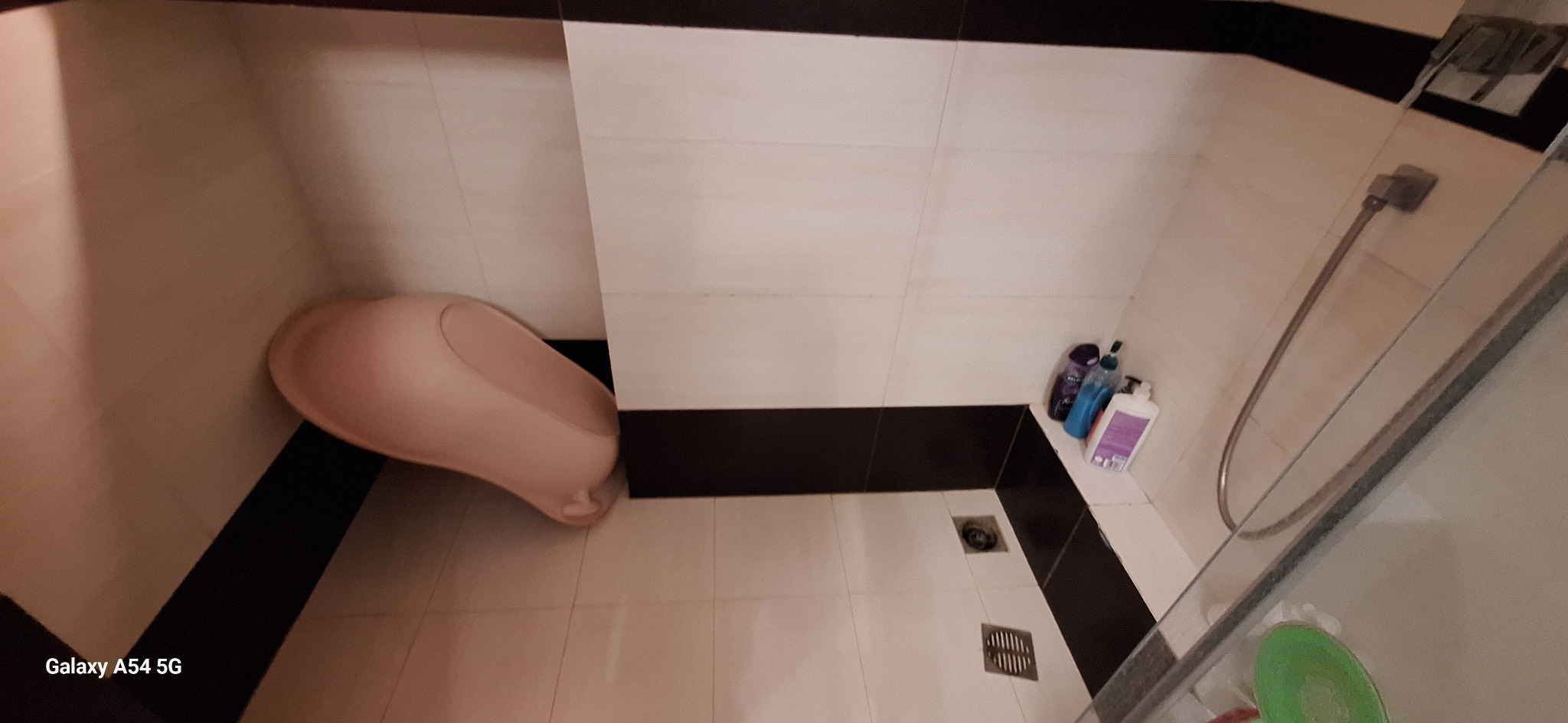 Wykończona łazienka z beżowymi i białymi płytkami, czarnym pasem dekoracyjnym, brodzikiem i kabiną prysznicową. Widoczne kosmetyki i odpływ liniowy.