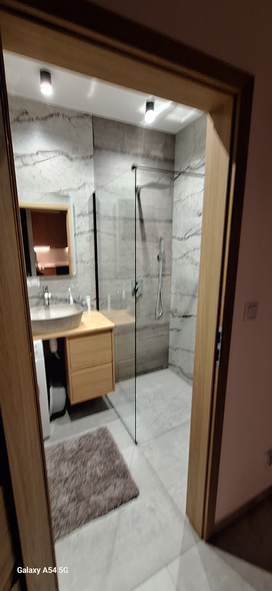 Nowoczesna łazienka z kabiną prysznicową typu walk-in, umywalką nablatową i szafką z jasnego drewna, wykończona płytkami imitującymi marmur.