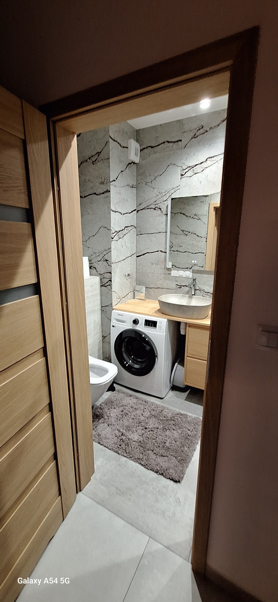 Nowoczesna łazienka z pralką pod blatem umywalki, szare płytki imitujące marmur, drewniane drzwi i szary dywanik. Widok zza otwartych drzwi.