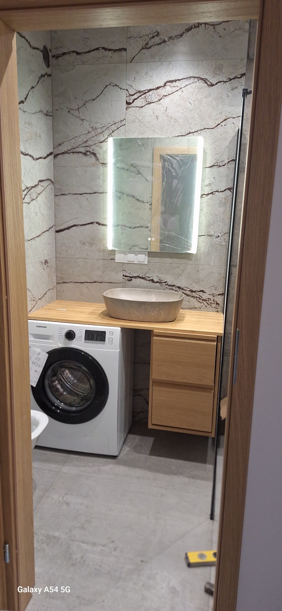 Nowoczesna łazienka z marmurowymi płytkami, podświetlanym lustrem, umywalką nablatową i pralką pod blatem. Minimalistyczny design z drewnianymi akcentami.