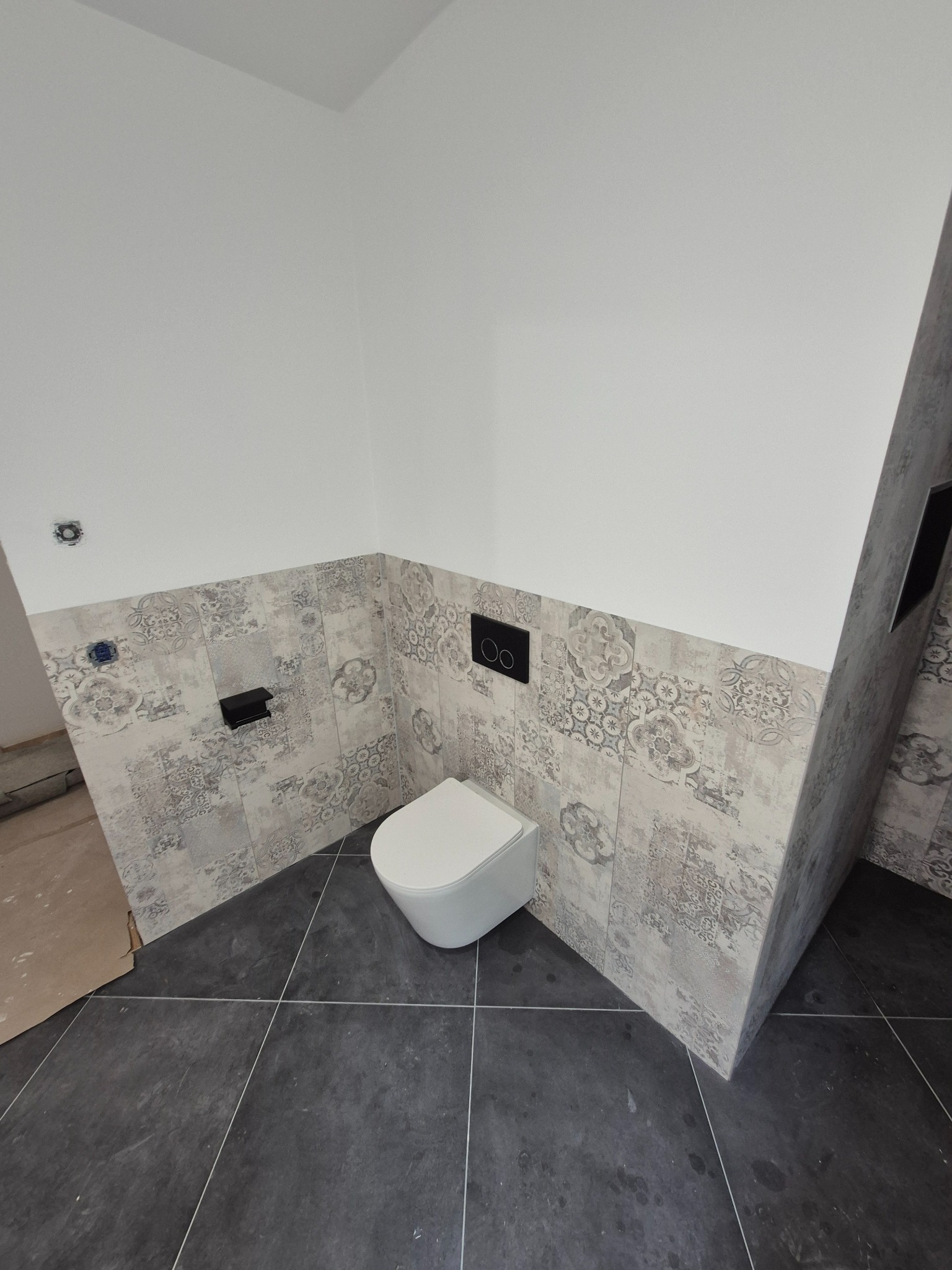 Nowoczesna toaleta w łazience z szaro-białymi płytkami dekoracyjnymi na ścianach i ciemną podłogą. Widoczne elementy instalacji i częściowo wykończona ściana.