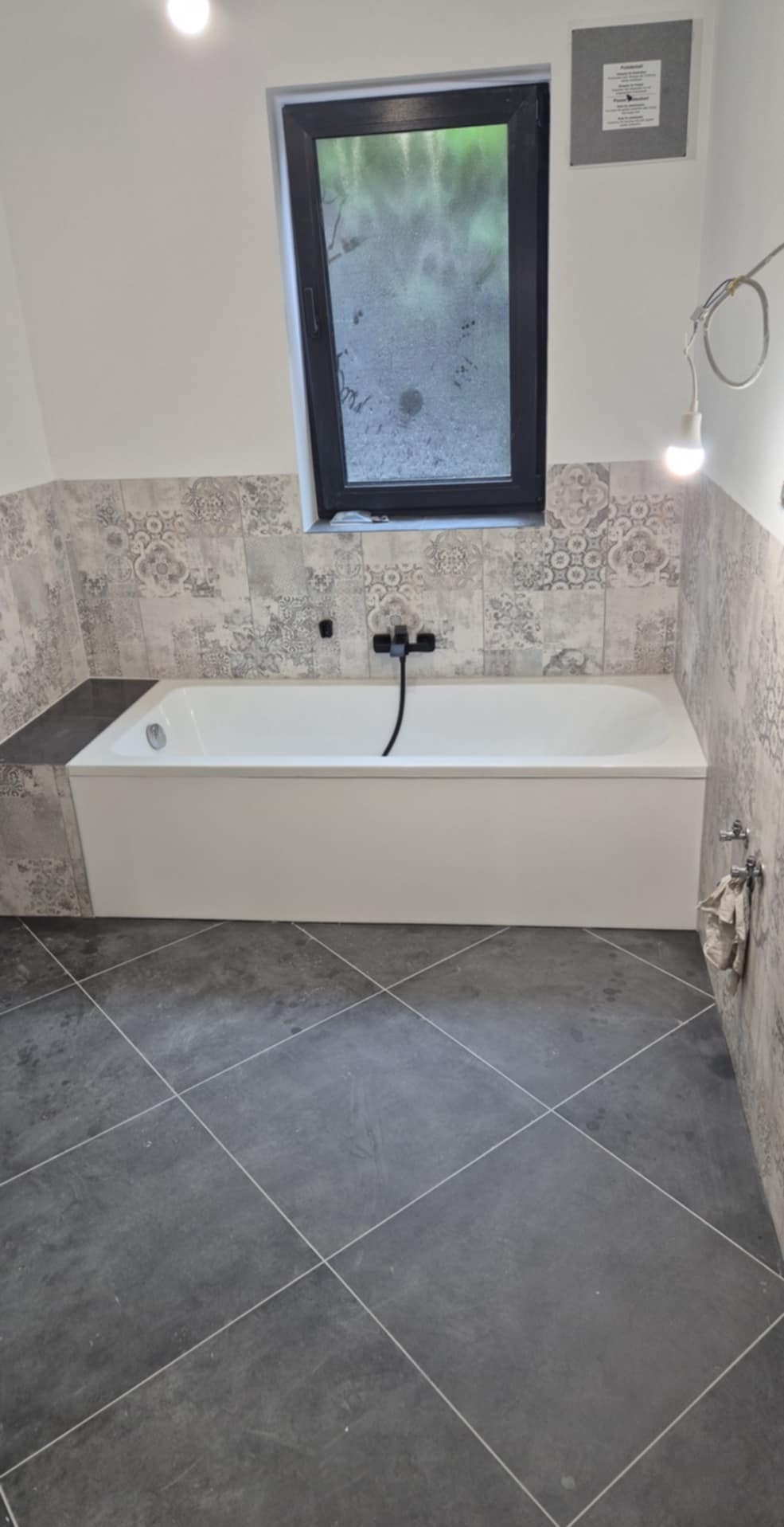 Wykończona łazienka z białą wanną, czarną armaturą i szaro-wzorzystymi płytkami na ścianach. Podłoga wyłożona ciemnoszarą terakotą w romby. Okno z czarną ramą.